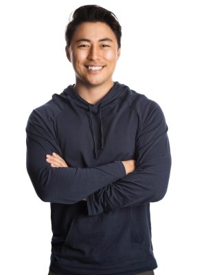 An attractive man in his 20s standing against a white background wearing a blue hood shirt smiling, looking at camera.